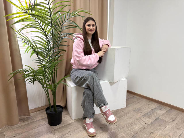 Lana Jenkin está sentada em um cubo branco, inclinada ligeiramente para frente, vestindo uma camisa rosa, calças cinzas e tênis rosa. Ela tem cabelos longos e pretos, e uma planta verde em um vaso está à sua esquerda..