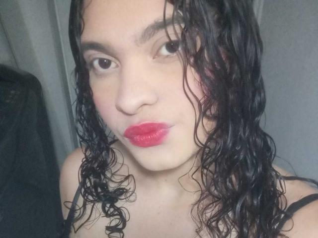Gros plan de NaughtyCare, avec des cheveux noirs bouclés et mouillés, une peau pâle et un rouge à lèvres rouge vif. Elle regarde directement l'appareil photo avec un léger sourire, et porte une bretelle noire. Incroyablement sexy.