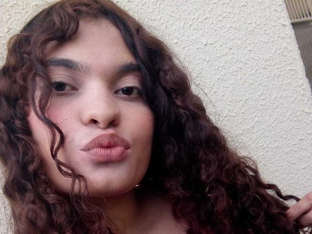 Nahaufnahme von NaughtyCare mit langen, lockigen braunen Haaren, vollen Lippen, die zu einem Kuss geformt sind, und dicken Augenbrauen. Helle Hautfarbe, leicht gerötet. Neutraler Hintergrund, dezente Schattierungen. Verdammt sexyer Look.