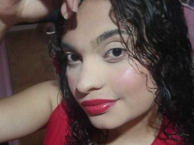 Primer plano de NaughtyCare con cabello negro rizado y húmedo, lápiz labial rojo y una ligera sonrisa. Ella lleva una blusa roja, con la mano levantada detrás de la cabeza. Su piel brilla bajo la luz intensa..