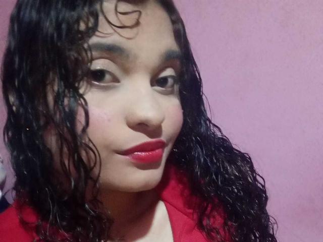 Close-up de NaughtyCare, uma mulher com cabelo preto encaracolado e molhado, pele clara e batom vermelho. Ela está vestindo uma jaqueta vermelha, com uma expressão sedutora, olhando diretamente para a câmera..