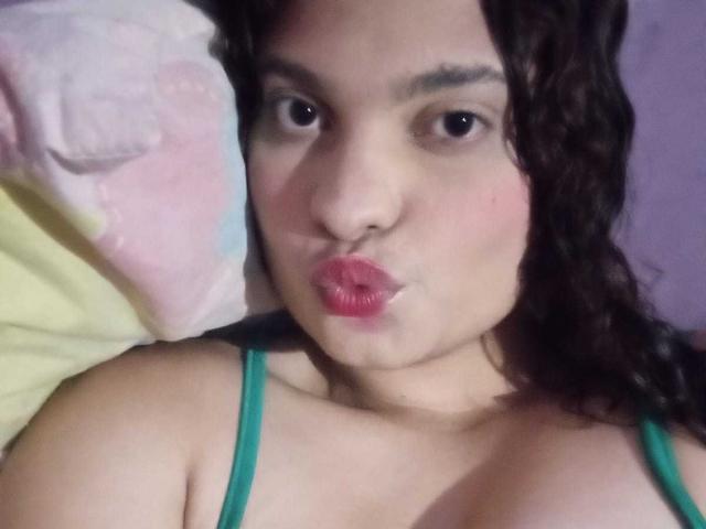 Close-up of NaughtyCare, a chubby woman with dark curly hair, pouting her red lips. She wears a green strap top, her dark eyes looking directly at the camera. Her chest is slightly visible.