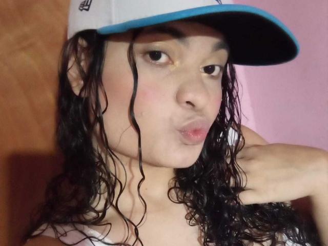 Close-up of NaughtyCare, a young woman with wet, wavy black hair, wearing a blue and white cap, puckering her lips in a pout. Her skin is light, and she's topless. Fucking sexy.