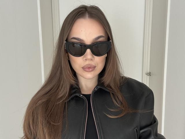 MirraBellle tiene el cabello largo y castaño, usa gafas de sol negras y una chaqueta negra con costuras blancas. Ella está mirando directamente a la cámara con una expresión neutra, transmitiendo una sutil tensión sexual..