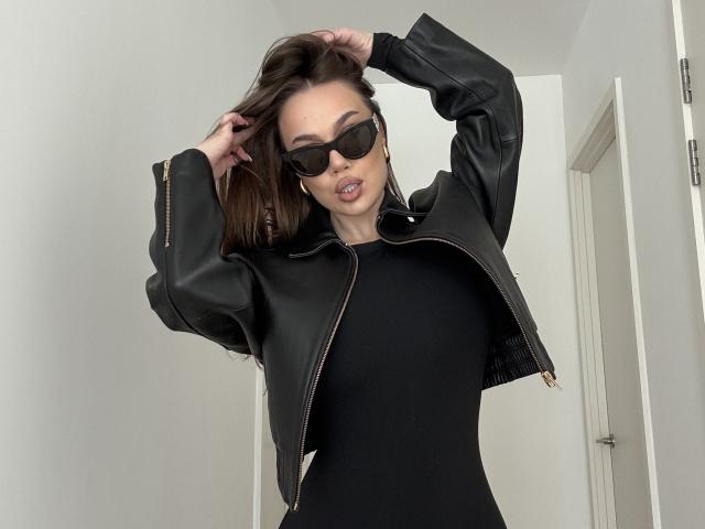 MirraBellle, een slanke vrouw met donker haar, draagt een zwarte zonnebril, een zwart kort jasje en een zwart topje. Ze staat met armen omhoog, en ze ziet er zelfverzekerd uit. Witte achtergrond, deur aan de rechterkant..