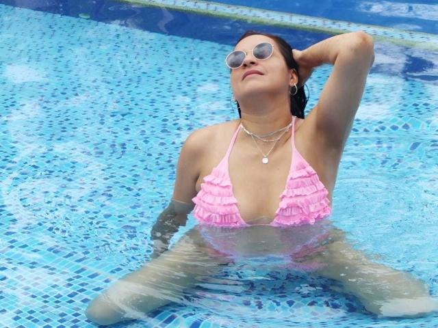 AliceDuboiss in a bright pink frilled bikini, kneeling in a blue-tiled pool, arm behind head, wearing round sunglasses, wet hair, necklace, water droplets on skin, relaxed expression, sunlight.