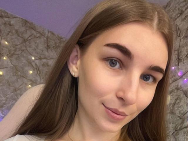 Autoportrait en gros plan de UBabyGilly, avec une peau claire, des yeux bleus et de longs cheveux bruns. Elle sourit légèrement, avec un maquillage discret et de petites boucles d'oreilles. Fond de mur en pierre avec des guirlandes lumineuses violettes et jaunes. Tellement mignon..