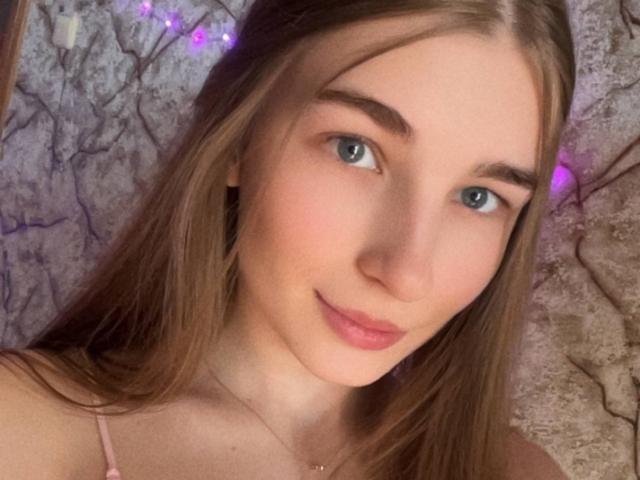 Nahaufnahme von UBabyGilly, einer jungen Frau mit heller Haut, blauen Augen und langen braunen Haaren, die leicht lächelt. Sie hat kleine Brüste und trägt ein dünnes, pinkes Top mit Trägern, vor einer strukturierten Wand..