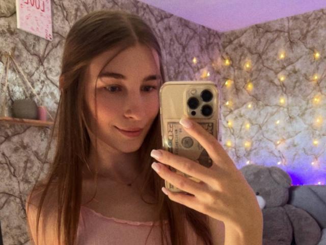 UBabyGilly, con el pelo largo y castaño, se toma un selfie frente al espejo con un iPhone, vistiendo una camiseta rosa. Luces de hadas cálidas y una pared con un diseño en el fondo. Absolutamente lindo y sexy..