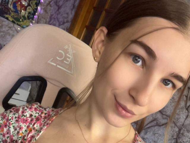 Autoportrait en gros plan de UBabyGilly, une femme à la peau claire, aux yeux bleus, aux cheveux bruns, et avec un tatouage d'un triangle blanc sur elle. Épaule gauche. Elle porte un haut à motifs floraux et sourit légèrement. L'arrière-plan est flou..