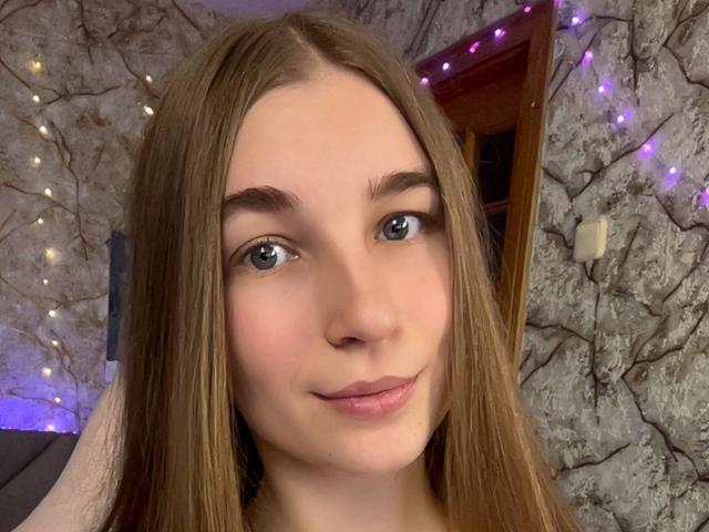 Close-up of UBabyGilly, a young woman with fair skin, long brown hair, and blue eyes, smiling slightly. Background has stone wall with purple and white fairy lights. Fucks look at her.