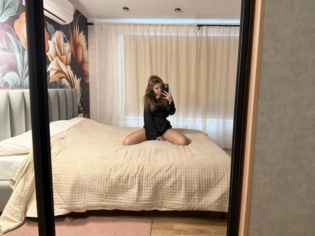 NiksaLove está arrodillada sobre una cama con una colcha blanca, vistiendo una camiseta negra y sin pantalones, y se está tomando una foto con el espejo. Está mirando a la cámara con una expresión seductora. Increíblemente sexy..