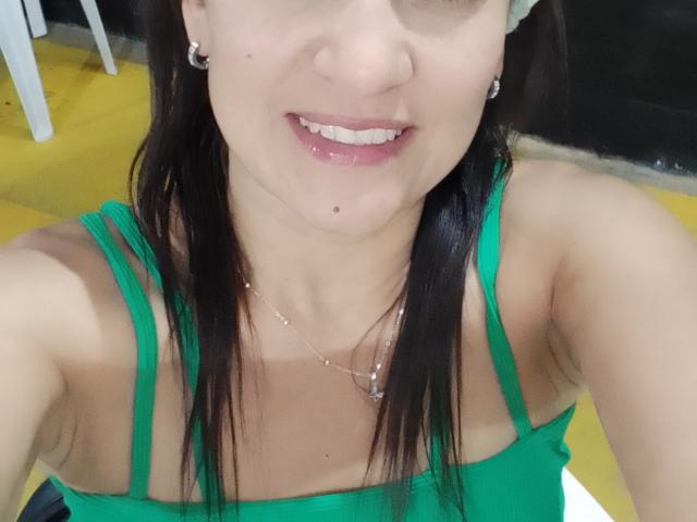 Selfie de perto de SolaraDubois com cabelo preto, blusa verde com alças finas, sorrindo com lábios rosados e brilhantes, visíveis. decote, colar pequeno, chão amarelo ao fundo. Atmosfera muito sensual, provocante e excitante..