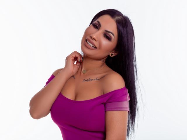 FragileHeidi, com longos cabelos pretos, vestindo uma blusa roxa justa, com um ombro de fora, mostrando o colo, sorrindo, com a mão perto do pescoço, tatuagem no peito, fundo branco, aparência sexy, pose confiante..