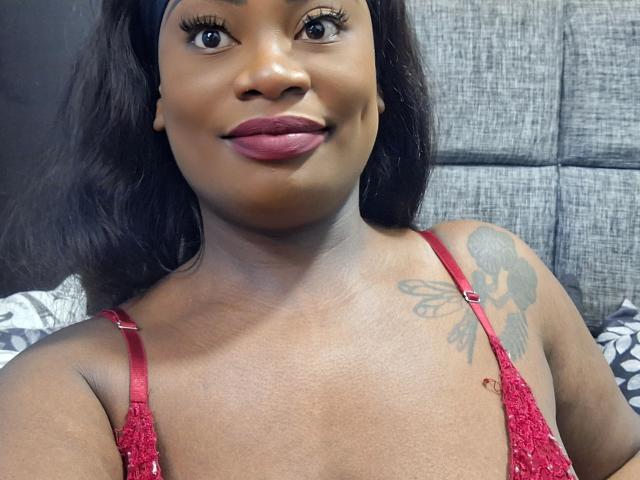 Primo piano di Alissa Jordan con la pelle scura, che indossa una top rossa con paillettes, mostrando un tatuaggio sulla spalla destra. Ha lunghi capelli neri, rossetto rosso e un leggero sorriso. Incredibilmente sexy..