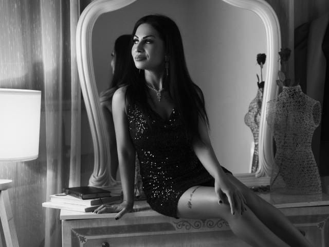 Schwarz-weißes Foto von FragileHeidi, die auf einem Schminktisch sitzt, ein glitzerndes Kleid trägt und langes, dunkles Haar hat. beleuchteter Spiegel, Mannequins im Hintergrund, sexuelle Pose, selbstbewusster Gesichtsausdruck, weiches Licht.