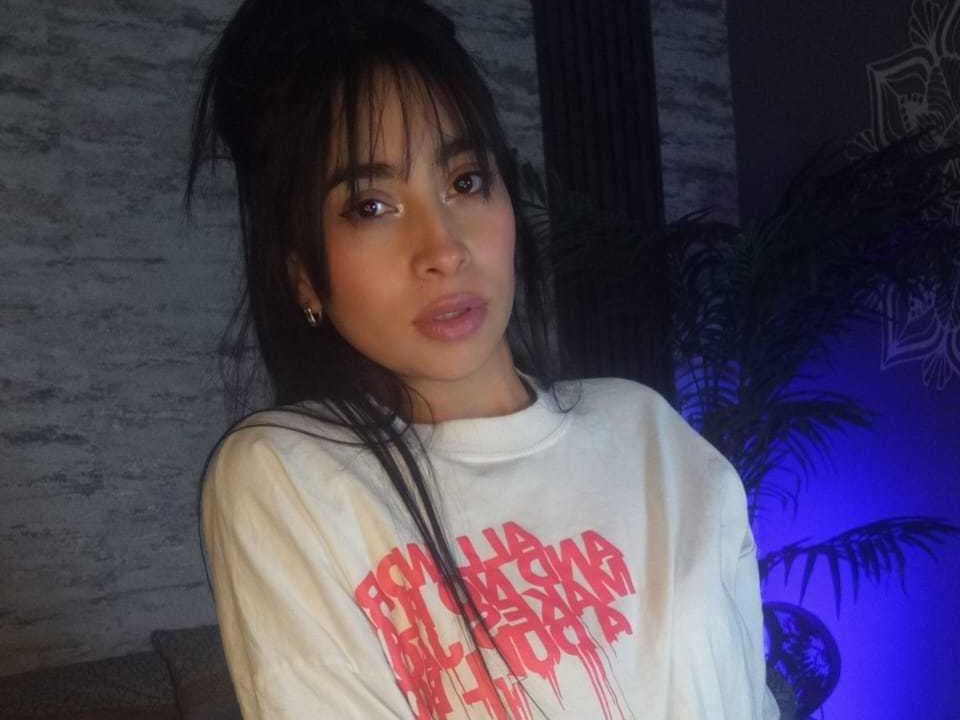 Photograph of SweetZoeZ, Asian woman with long black hair, wearing a white sweatshirt with red "PILLOSTAR PILLOSTAR" text. She gazes directly at the camera with a slightly seductive expression. Blue light in background.