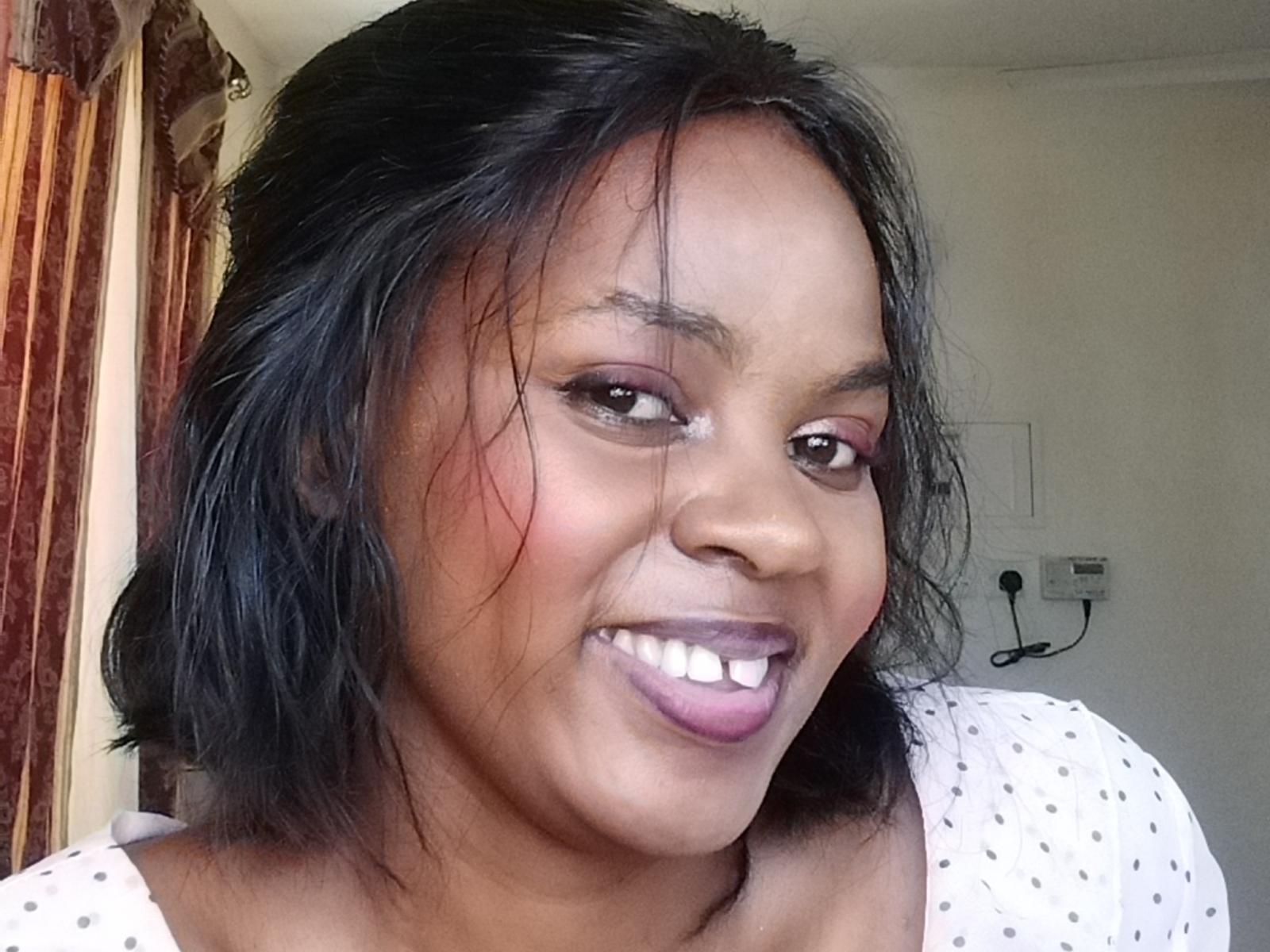 Close-up photo of KelishaLise with medium-dark skin, black shoulder-length hair, smiling with visible teeth, purple lipstick, wearing a white polka dot top, slight redness on right cheek, casual pose.
