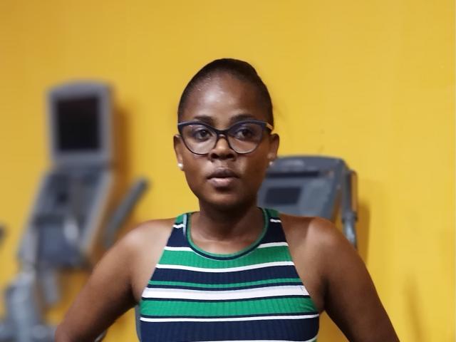 Fotografía de KelishaLise con piel oscura, cabello corto y gafas, vistiendo una camiseta sin mangas a rayas verdes y blancas, de pie. Contra un fondo amarillo brillante, con equipo borroso detrás de ella. Expresión muy seria..