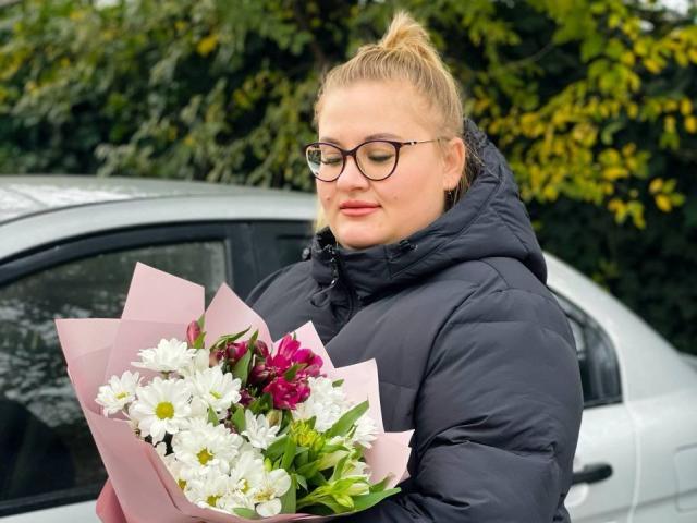 RadaRori mit Brille, blonden Haaren zu einem Dutt gebunden, trägt eine schwarze, gesteppte Jacke und hält einen Strauß aus weißen und roten Blumen. Stehend neben einem silbernen Auto, mit einem grünen, blättrigen Hintergrund..