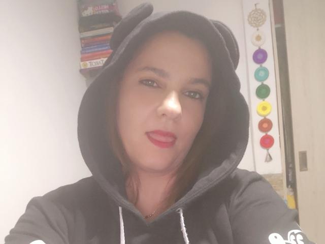 Amelia Wilson, vestida con una sudadera negra con capucha y orejas de oso, piel clara, lápiz labial rojo y ojos marrones, posando con una boca ligeramente abierta. Cuentas de chakra y libros en el fondo..