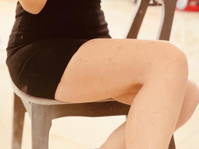 EllenVinny zit op een stoel, gekleed in een strak zwart jurkje, met gekruiste benen, waarbij haar gladde, bleke huid te zien is. Haar dijen zijn opvallend, wat een uitdagende en aantrekkelijke uitstraling creëert. De stoel heeft een donkerbruine afwerking..