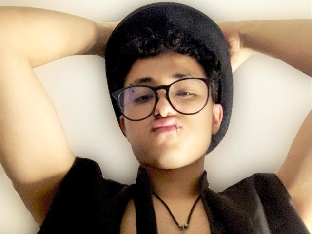 Adoni Baskerville, con cabello negro corto y gafas, con los brazos levantados detrás de la cabeza, vistiendo una camiseta negra y un collar, ligeramente... ojos entrecerrados, piel clara, foto casual con iluminación suave, pose relajada..