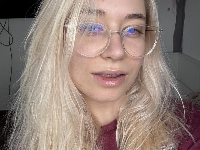 Nahaufnahme von BeckyThaissa mit zerzaustem blondem Haar, die runde Brillen mit blauen Reflexionen und ein burgunderfarbenes Hemd trägt. Sie hat den Mund leicht geöffnet, und die Beleuchtung betont ihre Lippen..