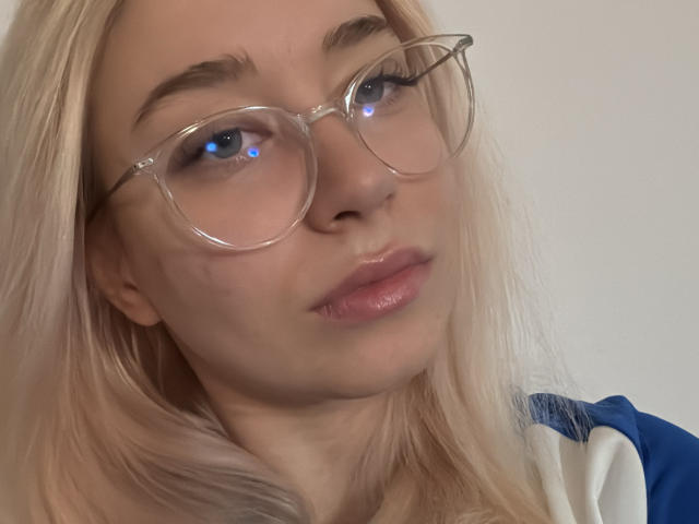 Nahaufnahme von BeckyThaissa mit platinblondem Haar, die randlose Brille, glänzende rosa Lippen und ein blau-weißes Outfit trägt. oben. Blaue Lichtreflexion in den Augen, leichte Narbe im Gesicht auf der linken Wange..
