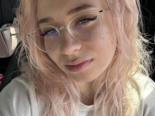 Photo en gros plan de BeckyThaissa, avec des cheveux blonds platine, portant des lunettes rondes avec du crayon noir pour les yeux, et une chemise blanche. Elle a une expression séduisante, avec les lèvres légèrement entrouvertes, et une tension sexuelle visible..