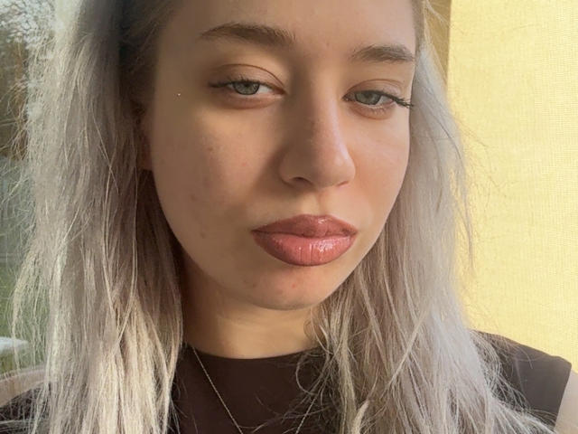 Close-up de BeckyThaissa, uma mulher de pele clara com cabelos longos e prateados, lábios vermelhos cheios e algumas sardas. Ela tem um olhar sedutor, maquiagem leve e uma expressão confiante, ligeiramente provocante. A luz do sol realça seu rosto..