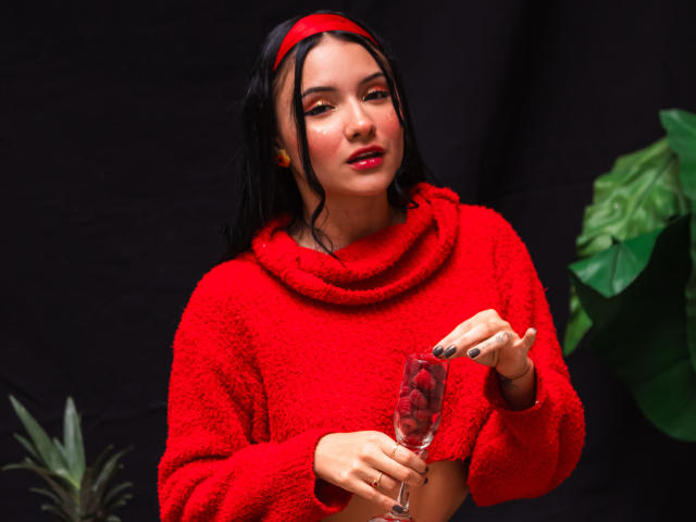 Ilustração digital de LizzeJoy com cabelo escuro, uma faixa vermelha e um suéter vermelho, segurando uma taça de vinho. Ela está olhando sedutoramente para a câmera, com uma planta ao fundo. Vermelhos e pretos vibrantes.