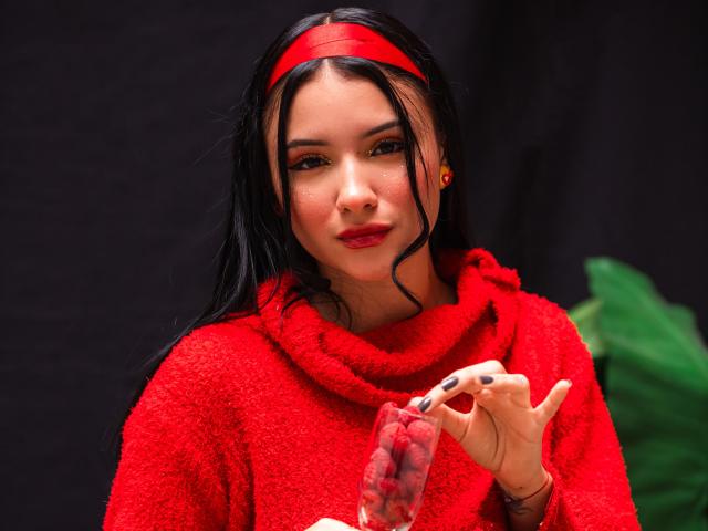 LizzieJoy mit schwarzen Haaren, einem roten Stirnband und einem roten Pullover, hält eine Packung Kondome in roter Verpackung nahe ihrem Gesicht. Schwarzer Hintergrund, ein grünes Kissen ist leicht sichtbar. Verführerischer Ausdruck, sexuelle Spannung..