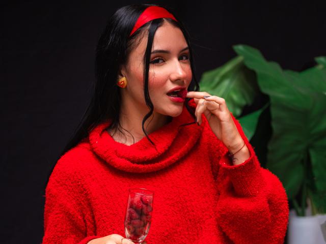 LizzieJoy, con i capelli neri, una fascia rossa e un maglione rosso, sta mordendo un lecca-lecca rosso, tiene in mano un bicchiere con una bevanda, sfondo scuro. sfondo, pianta verde, sguardo intenso, tensione sessuale, colori rossi vivaci.