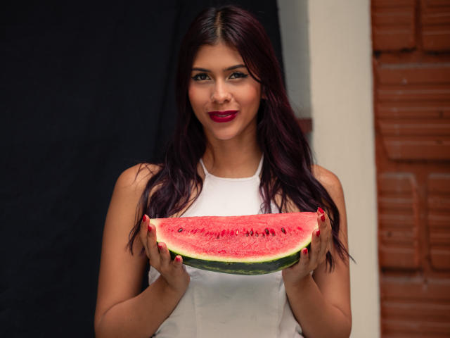 DanikaSerov, une femme latina séduisante avec de longs cheveux noirs et du rouge à lèvres, tient une tranche juteuse de pastèque, sa peau haut sans manches mettant en valeur sa silhouette élancée et sa poitrine de taille moyenne..