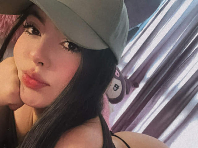 Close-up van CamillaRiley, die een groene muts draagt, zwart haar heeft en rode lippenstift, en haar wang op haar hand rust. De achtergrond heeft gestreepte lichtpatronen en een pluche speelgoed. Ongelooflijk sexy, een schattig gezicht..