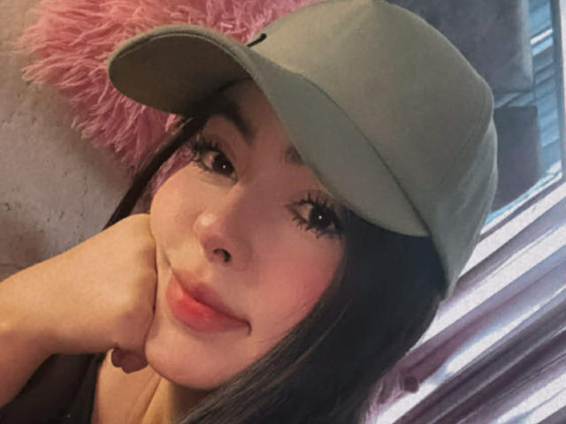 Close-up of CamillaRiley, an Asian woman with long black hair, wearing a green cap, resting her hand on her cheek, smiling with red lipstick, natural light from the right, pink fluffy pillow.