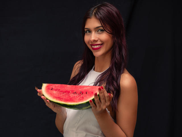 Fotografia de Danika Serov, uma mulher de cabelos escuros com batom vermelho, segurando uma fatia de melancia, vestindo uma blusa branca sem mangas. fundo preto, o vermelho e o verde da melancia contrastando com a sua pele, sorrindo de forma sedutora..