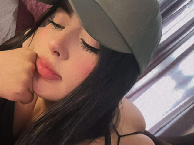 Fotografía de primer plano de Camilla Riley, una joven con cabello largo y negro, que lleva un gorro verde, con los ojos cerrados y apoyando la mejilla. en su mano, labios rosados ligeramente entreabiertos, sexy y relajada.