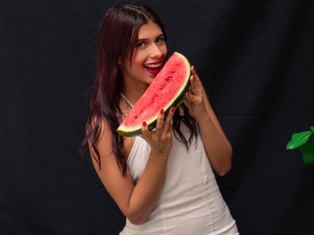DanikaSerov, a young woman with long dark hair, wearing a white tank top, holds a juicy, red watermelon slice with both hands. She's smiling with red lips, looking directly at the camera.