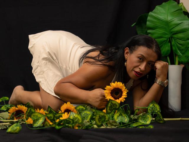 SoffieMature, eine kurvige Frau mit dunkler Haut und langen schwarzen Haaren, liegt auf dem Bauch inmitten von Sonnenblumen und grünen Blättern. Sie trägt einen weißen Rock und blickt direkt in die Kamera, mit einem verführerischen Lächeln..