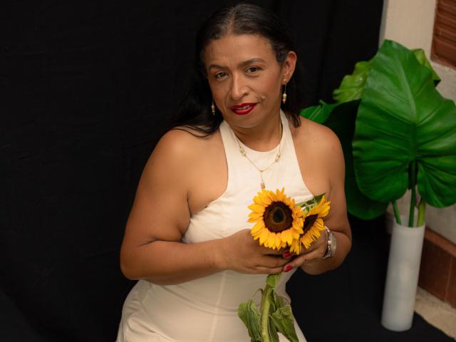 SoffieMature, con la pelle abbronzata e i capelli neri, indossa un abito bianco con spalline e tiene un girasole vicino al petto. Ha il rossetto rosso e degli orecchini, e sorride leggermente. Sfondo nero, pianta verde..