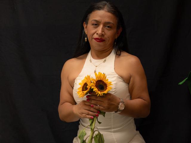 SoffieMature, una donna più matura con i capelli scuri, tiene in mano un girasole giallo brillante, indossando un abito bianco con spalline e unghie rosa. rossetto rosso, orecchini dorati e un orologio. Sfondo nero. Tensione sessuale percepibile dallo sguardo..