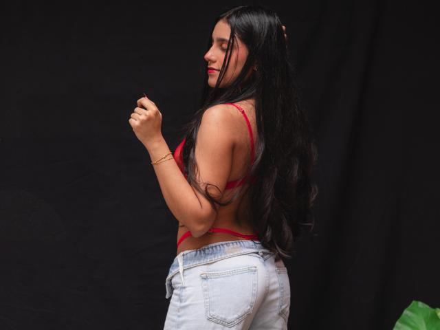 TiffannyRose est en silhouette de profil, avec de longs cheveux noirs, un soutien-gorge rouge, un jean blanc, et le poing droit levé. Fond noir. Femme séduisante, aux formes généreuses, peau bronzée, rouge à lèvres rouge, pose confiante, dos légèrement arqué, allure coquette..