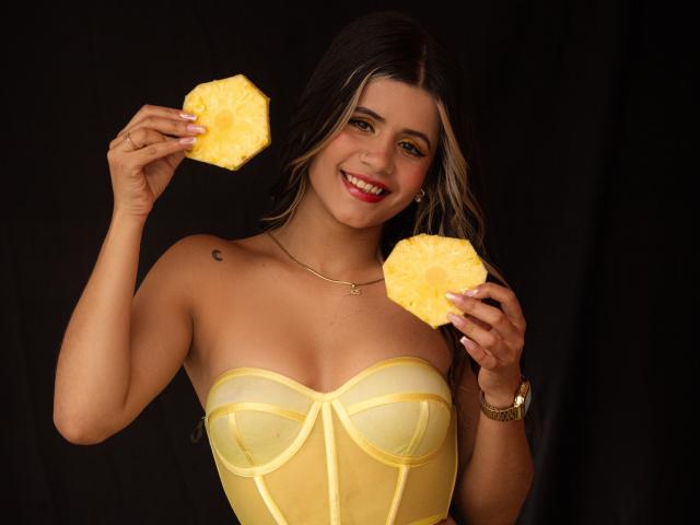 SheylaSpooner, a hot Latina woman with long dark hair, wearing a yellow strapless corset, holding two yellow mango pieces, smiling, showing a sexy pose, black background, visible tan lines.