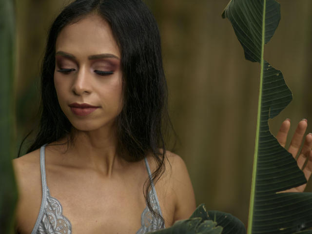 Fotografía de Aliicia Smith con cabello largo y negro, vistiendo una blusa de encaje plateada, mirando hacia abajo con los ojos cerrados. Sosteniendo una hoja verde grande, húmeda y brillante. Tensión sexual reflejada en su expresión concentrada..