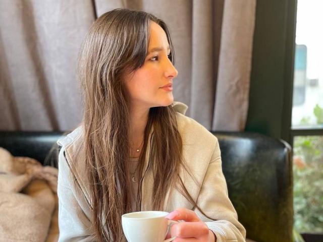 Omella, with long brown hair, sits in a black leather chair, holding a white cup. She gazes to the side with a slight smile. Grey curtains and sunlight in the background.