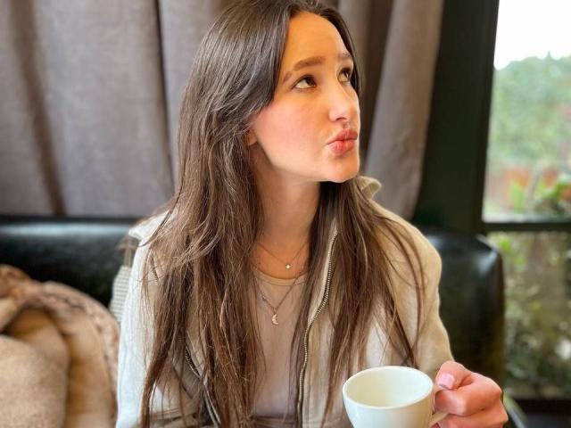 Omella, mit ihren langen, braunen Haaren, blickt mit leicht geöffnetem Mund nach oben und hält eine weiße Tasse. Sie trägt ein weißes Oberteil, eine Halskette und ihr Gesichtsausdruck verrät eine sanfte, sexuelle Anziehung..