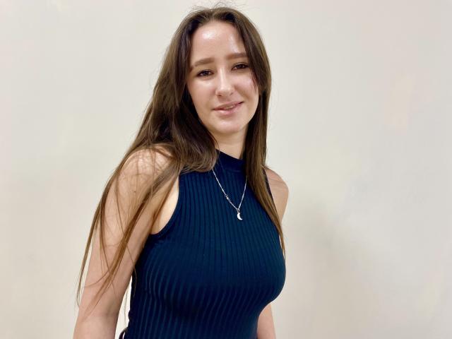 Omella tem cabelos castanhos longos, veste uma blusa preta canelada sem mangas, está sorrindo levemente e usa um colar. Sua expressão é calma, sua linguagem corporal está relaxada. A iluminação é forte, destacando suas características..
