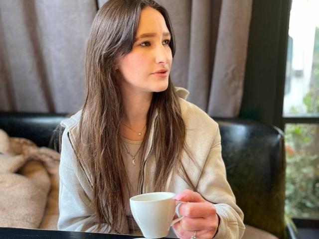 Omella, with long brown hair, sits holding a white cup, wearing a white shirt. She gazes to the side, showing a slight smirk. Her hand rests on the table. Fucking casual.